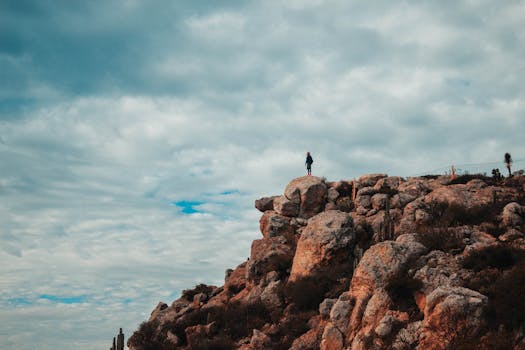 A person stands on a cliff amidst rock formations under a cloudy sky, symbolizing adventure and exploration.