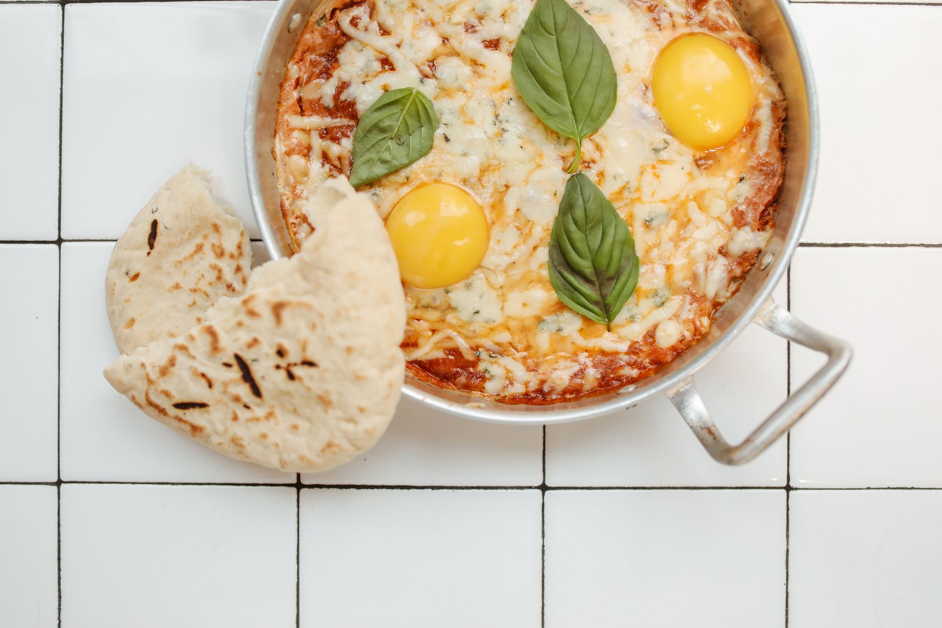 Appetizing shakshuka topped with basil leaves, served with pita on a tiled background.
