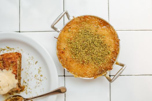 Top view of knafeh and baklava with creamy topping, garnished with pistachios on white tiles.