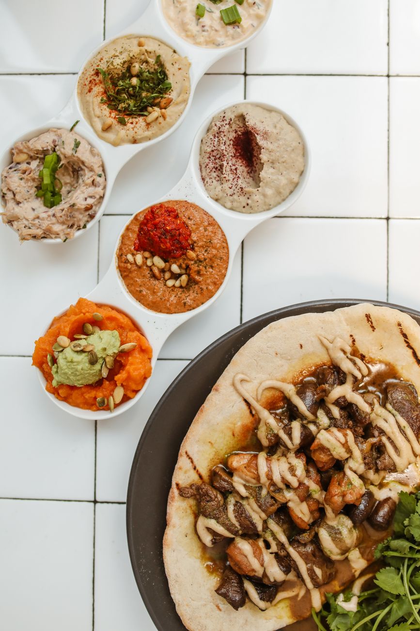 Three bowls of Middle Eastern spice pastes - reddish muhammara with visible walnut pieces, dark jammy acuka, and bright green chunky zhug - surrounded by fresh herbs, walnuts, and red peppers
