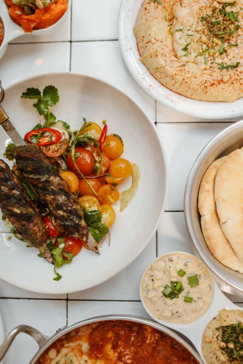 Free A delicious Mediterranean spread featuring kebab, hummus, tomatoes, and flatbread on a tiled table. Stock Photo