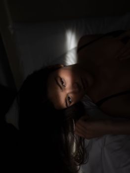 From above of young sensual female with long dark hair in lingerie lying on soft bed and looking at camera