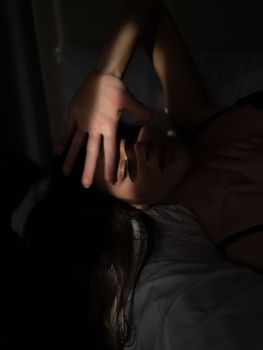 From above of feminine young lady with long dark hair in underwear lying on bed with closed eyes and covering face from sunlight with hand