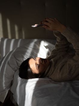 Self assured of young female with long dark hair lying on comfy bed and taking selfie on smartphone in sunny morning