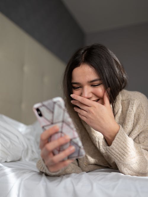 Cheerful young lady covering mouth with hand while reading funny message in smartphone lying on comfortable bed at home