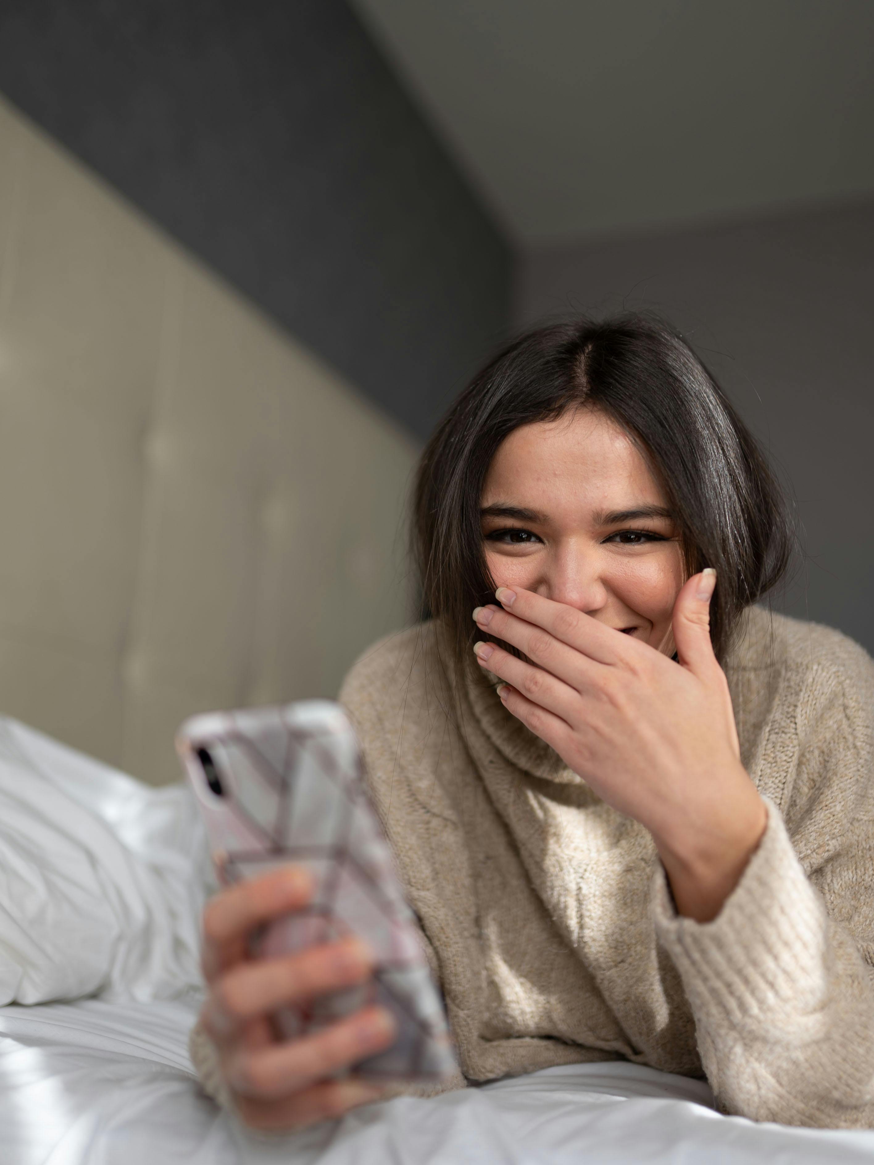 joyful young woman smiling while browsing smartphone on bed
