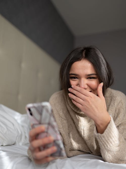 Joyful young woman smiling while browsing smartphone on bed