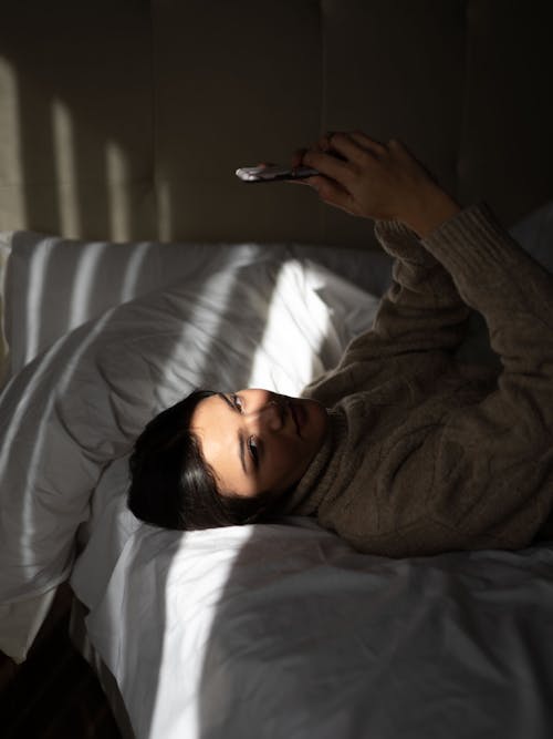 Side view of confident young female in warm sweater recreating on soft bed with smartphone in hands and looking at camera