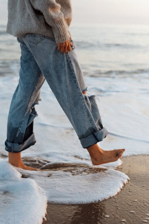 Side view of crop unrecognizable barefooted female tourist in stylish jeans and sweater walking on sandy beach washing by foamy waves of ocean during holidays