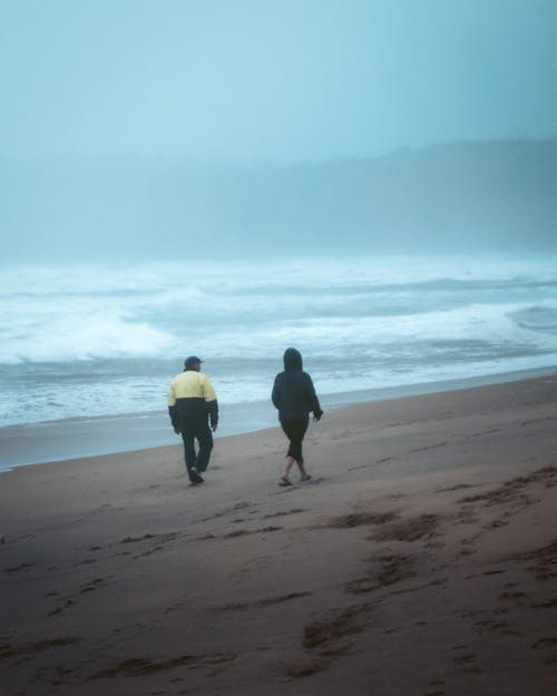 Unrecognizable couple walking on sandy coast near wavy sea under gloomy sky