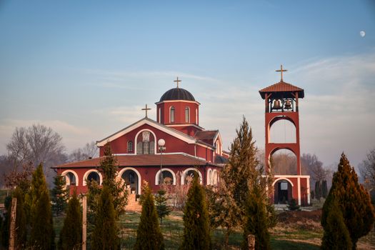 A stunning red chapel with bell tower surrounded by lush countryside under a clear sky.