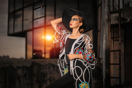 Gorgeous female with stylish makeup wearing elegant outfit and broad brim standing with hand on head at sunset