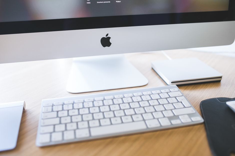 An iMac with the Apple logo on a wooden desk, an Apple wireless keyboard in front of it and a notebook to the side