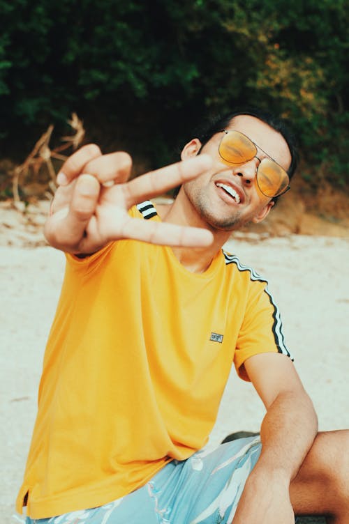 Young cheerful male in bright yellow T shirt smiling and looking at camera while giving V sign on beach