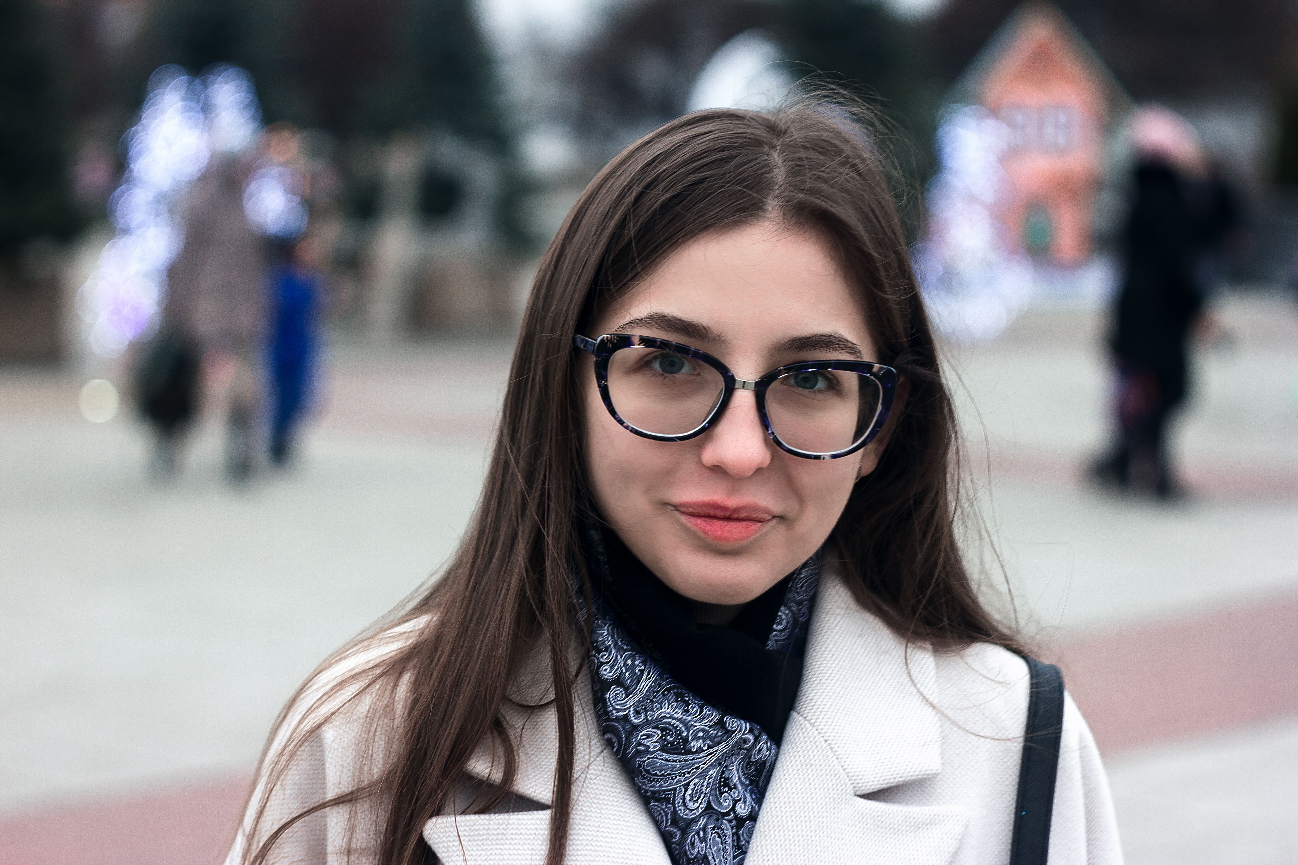 Young woman in glasses smiling outdoors, wearing a white coat and paisley scarf.