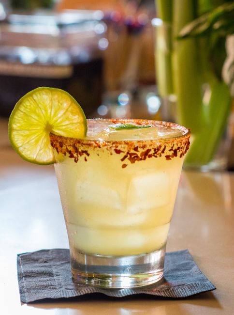 A pale gold margarita in a salt-rimmed rocks glass over ice, garnished with a lime wheel, next to a small dish of kosher salt and fresh limes.