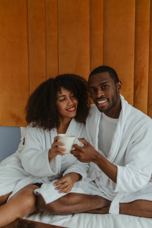 A Couple Sitting on the Bed While Wearing Bathrobe and Holding a