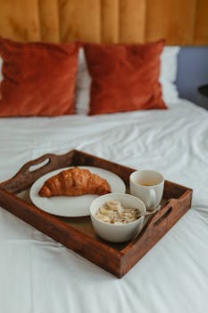 A wooden tray with a croissant, cereal, and coffee on a comfy bed with cozy pillows.