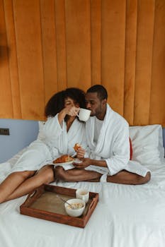Couple in bathrobes having breakfast in bed, sharing intimate moments indoors.