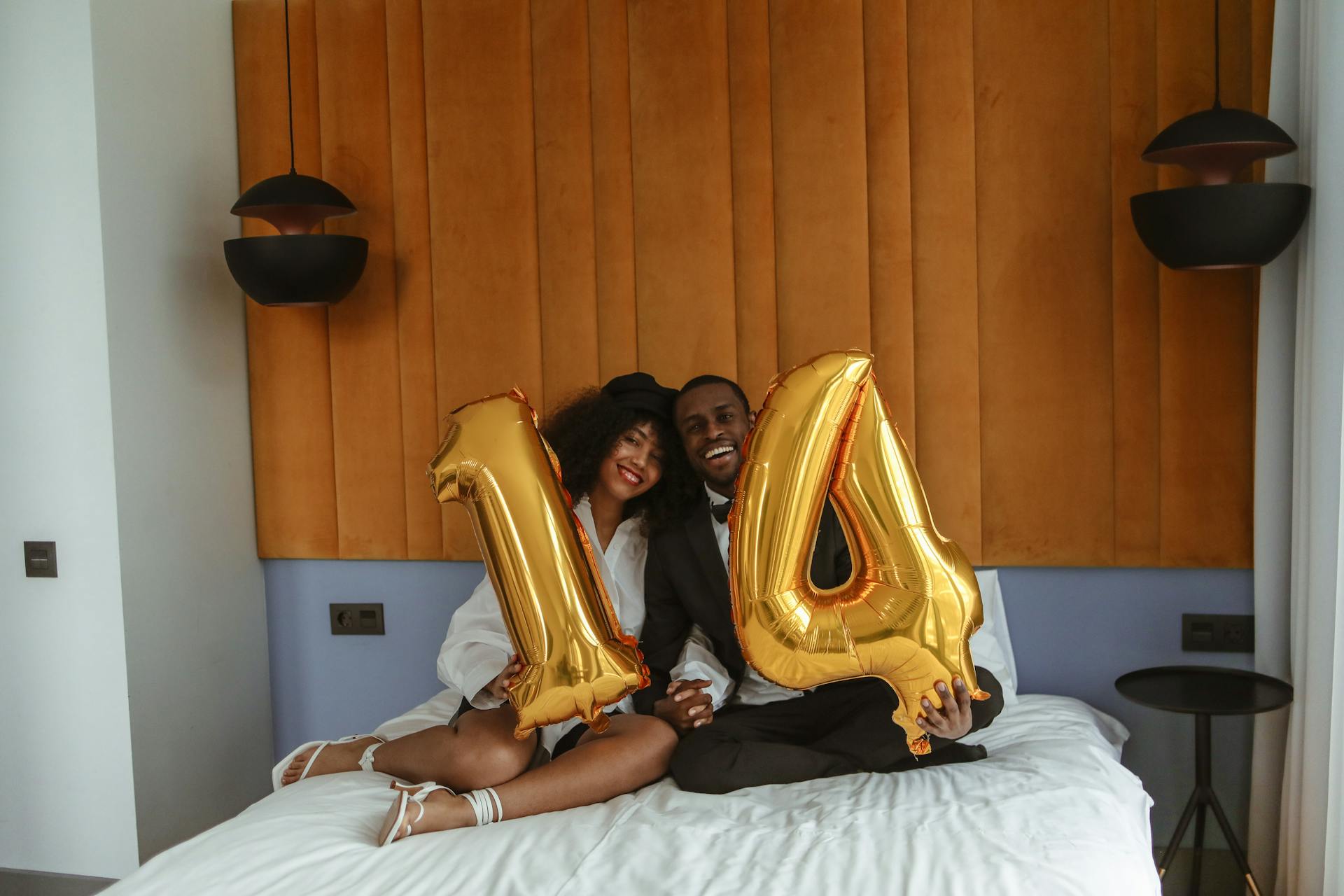 Smiling couple celebrating 14th anniversary with golden balloons on bed indoors.