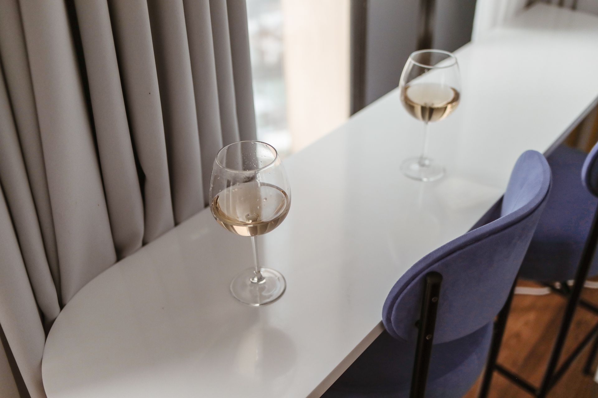 Two wine glasses filled with white wine on a sleek modern bar table with blue chairs indoors.