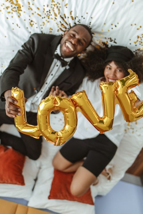 Free Smiling couple in formal attire holding love balloons on a bed with confetti, capturing a joyful and romantic moment. Stock Photo