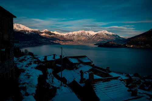 Free Small residential houses with snowy roofs located on shore of calm lake surrounded by rocky mountains against cloudy sunset sky in winter Stock Photo