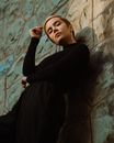 Low angle of young confident female in trendy black clothes standing in light place near shabby uneven surface while looking at camera