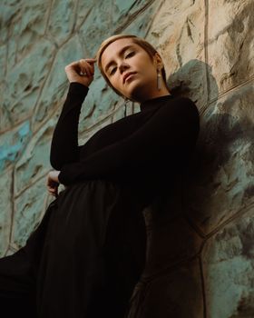 Low angle of young confident female in trendy black clothes standing in light place near shabby uneven surface while looking at camera