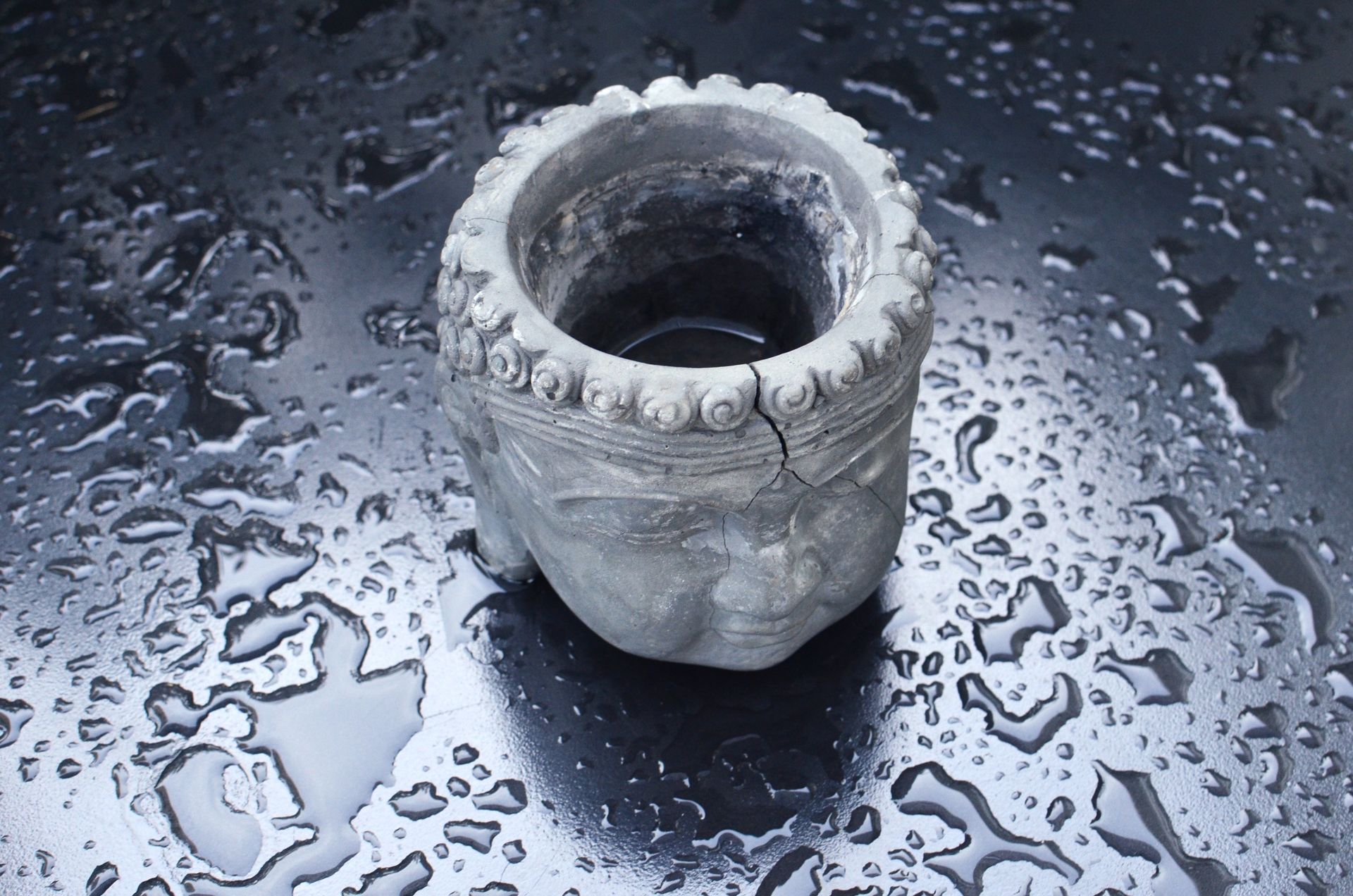 Ornate ceramic Buddha head planter on wet surface with waterdrops, showcasing texture and heritage craftsmanship.