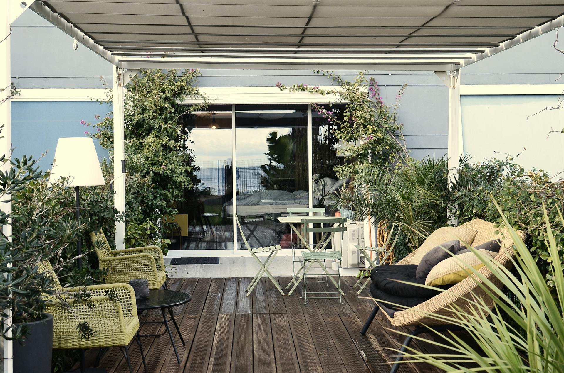 Comfortable stylish rattan armchairs with tables placed on wooden veranda of modern residential villa with lush exotic plants on sunny day