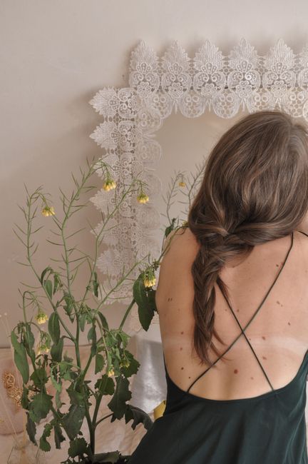 A woman with braided hair sits in front of a lace-adorned mirror and plant, creating an elegant atmosphere.
