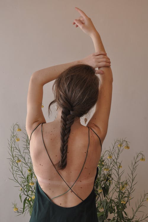Free Back view of unrecognizable female with pigtail standing near blooming plant with hand above head Stock Photo