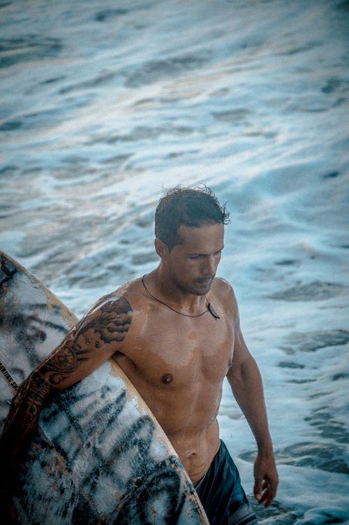 Free A shirtless man with a tattooed arm carrying a surfboard by the ocean. Stock Photo