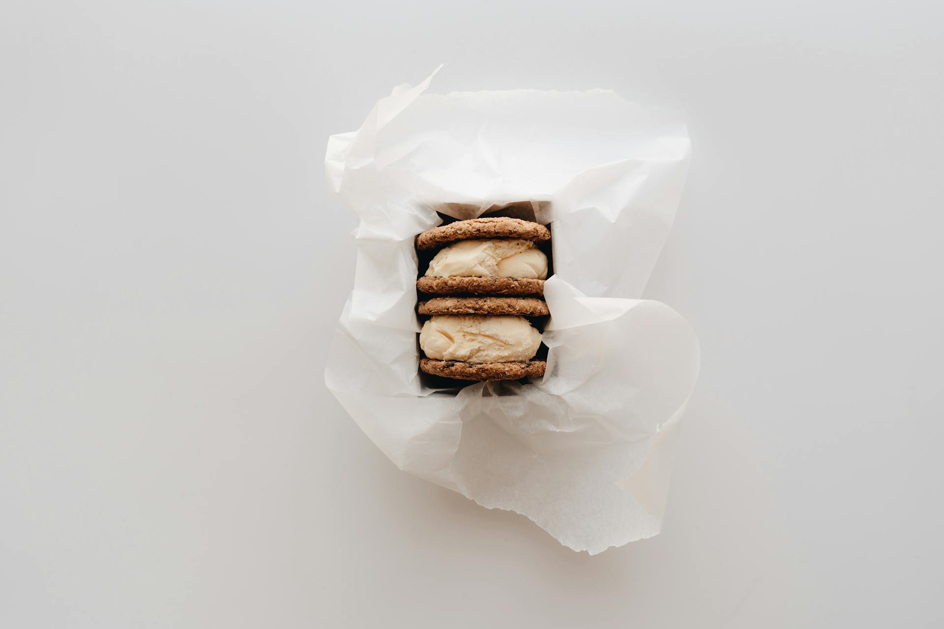 Three ice cream cookie sandwiches wrapped in wax paper on a white background.