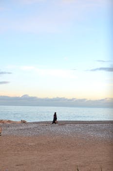 Single person walking along a serene beach during dusk with a calm ocean and expansive sky.