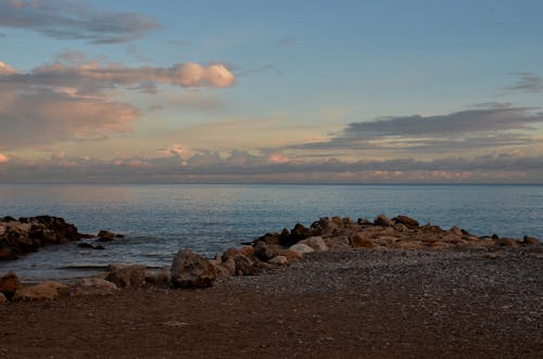 Picturesque view of rough coast with stones against rippled ocean under cloudy sky with horizon at sundown