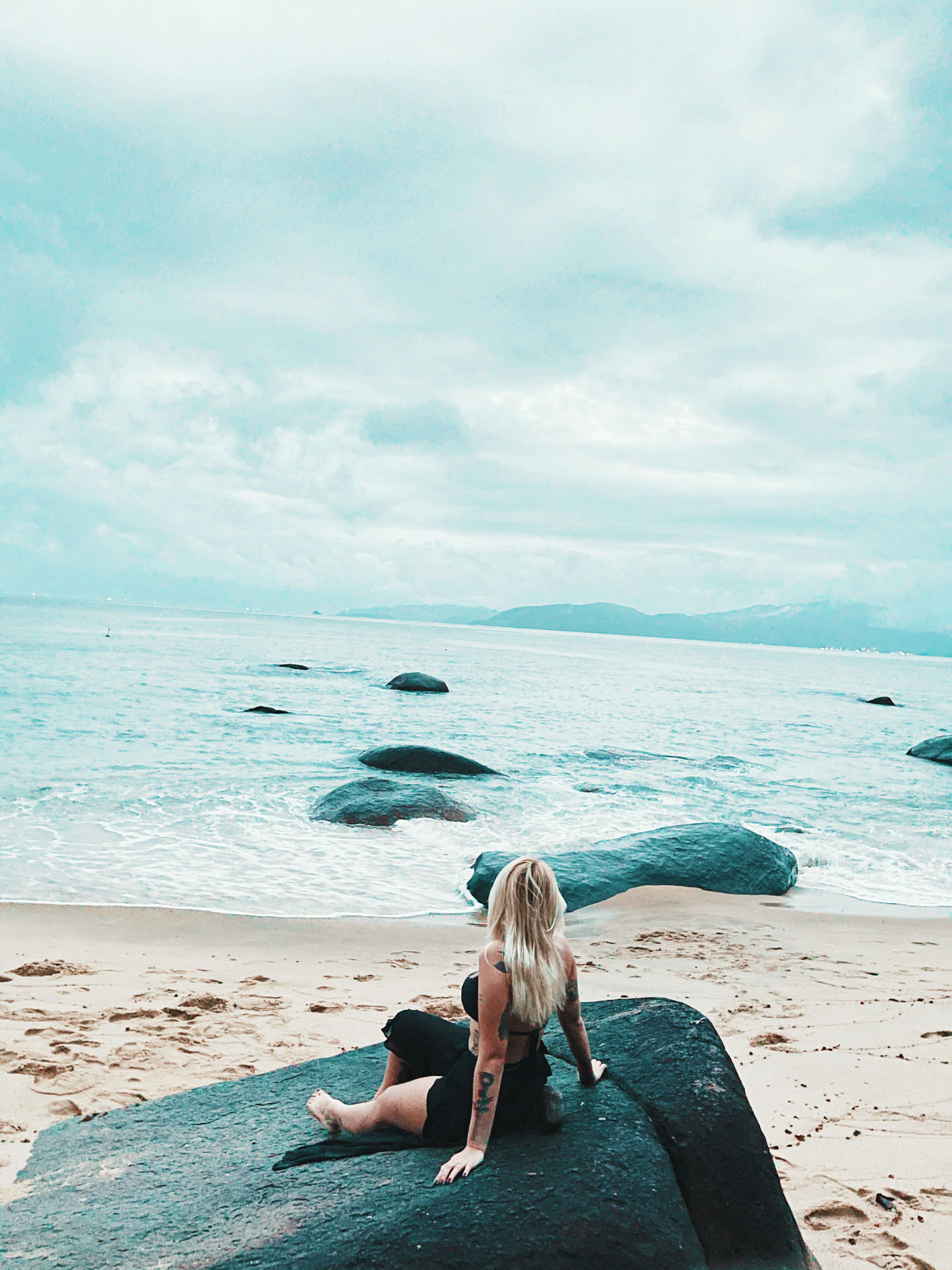 A young woman with blonde hair sits on a rock by the seashore in Conceição de Jacareí, Brazil, under a cloudy sky.