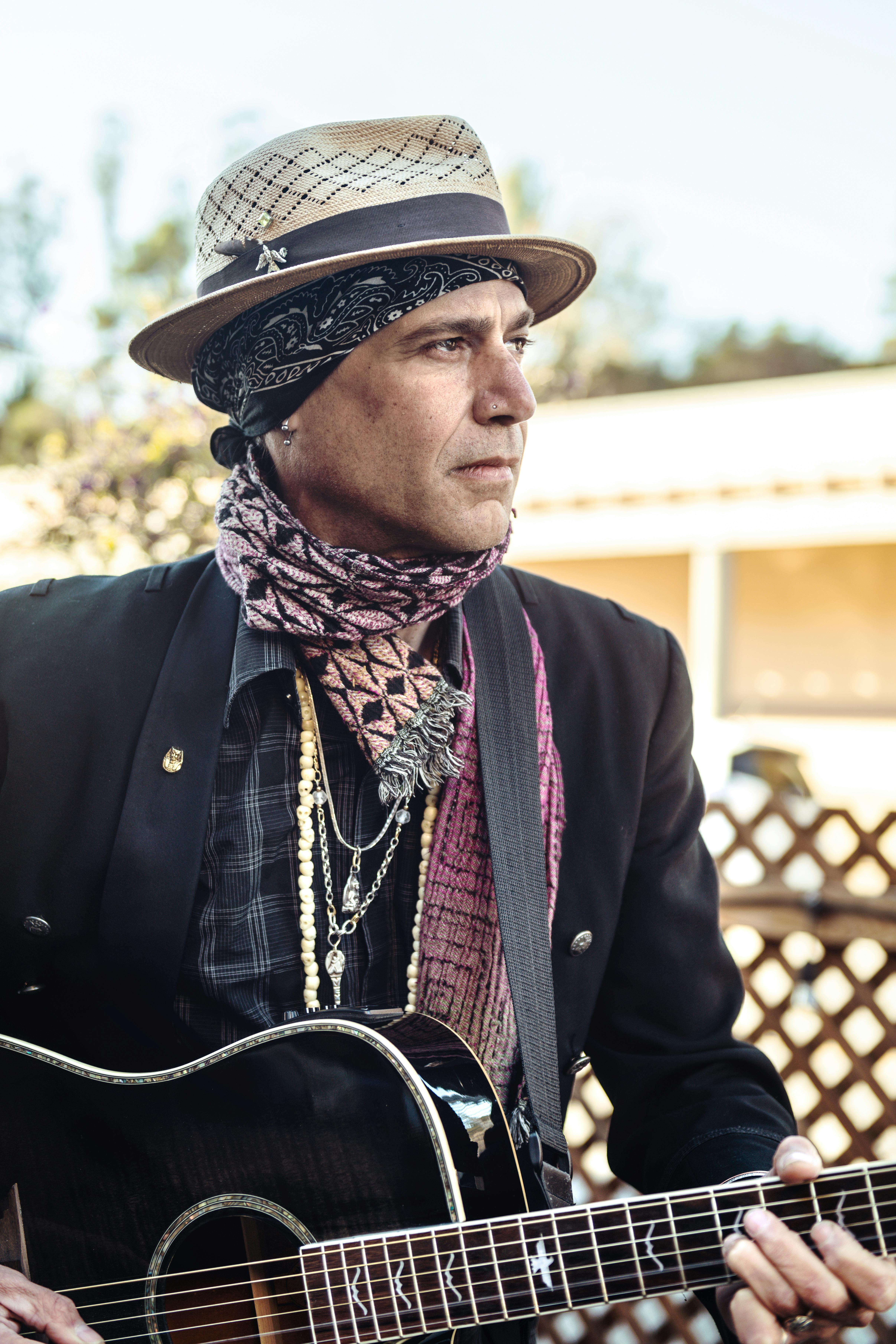 A stylish musician in Cambria, CA plays an acoustic guitar, embracing a bohemian look with a hat and scarf.