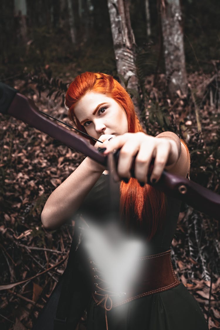 Serious Redhead Female Archer In Medieval Costume Hunting With Bow In Forest