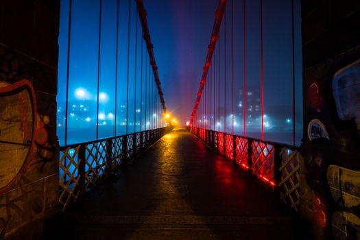 A neon-lit suspension bridge glowing in misty conditions during the night in Glasgow.