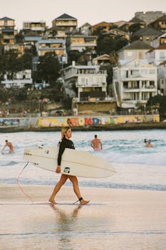 A surfer exits the ocean carrying a surfboard with urban residences in the background.
