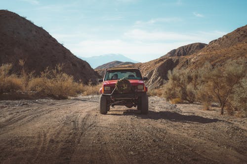 Red SUV with bullbar riding on rural roadway in nature among lush bushes and mountainous terrain against cloudy sky in countryside