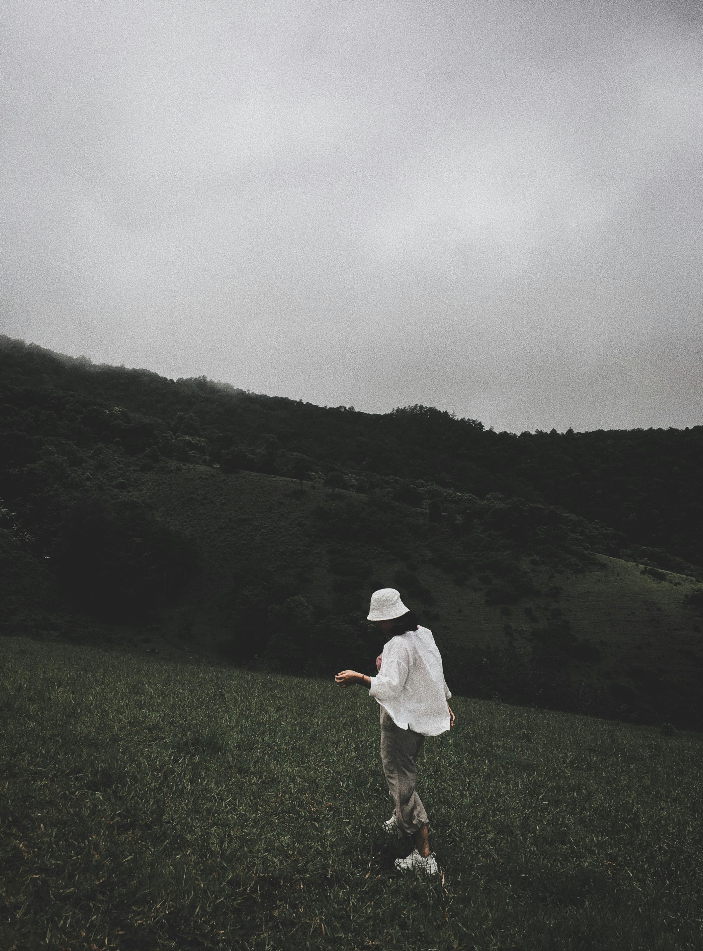 A solitary woman in a white hat walking through a lush green meadow under a cloudy sky.