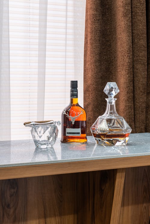 Transparent bottle and decanter with expensive alcoholic drink near glass with cigar on table in house
