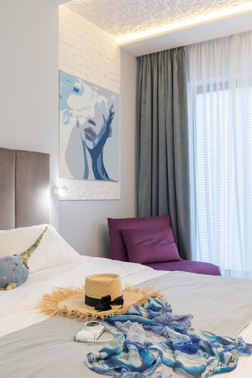 Modern hotel room interior with straw hat and photo camera on soft bed against curtains