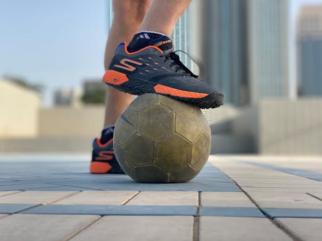 Close-up of a sneaker resting on a soccer ball in a modern urban landscape.