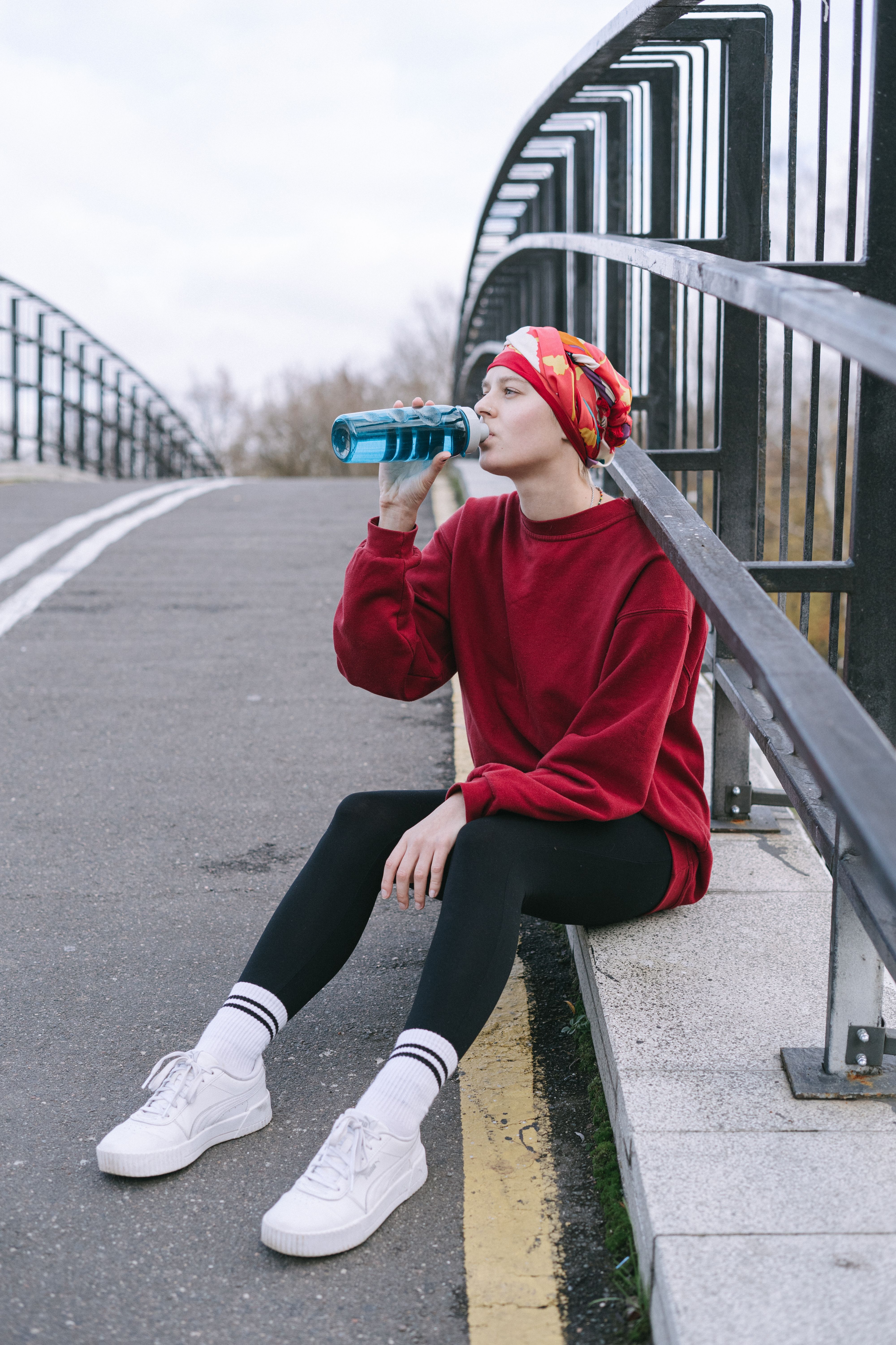 Casual young woman wearing a headscarf, sitting on a bridge, and drinking from a bottle.
