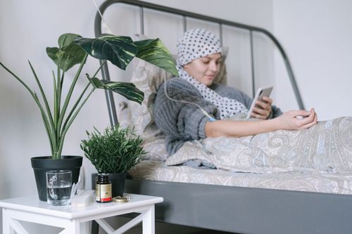 Free A woman undergoing cancer treatment relaxes in bed using her phone, surrounded by plants. Stock Photo