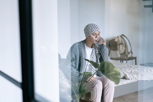Free Woman in headscarf sitting on bed indoors, drinking water, significant in cancer awareness and treatment support. Stock Photo